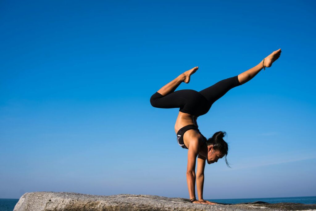 A woman performing a yoga handstand on a rock by the sea under a clear blue sky.